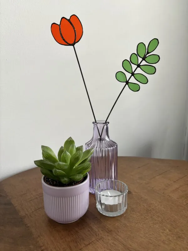 Tiffany glaskunst: oranje tulp in een vaasje met een 9-blad tak met ronde blaadjes, beide bevestigd op een steel.