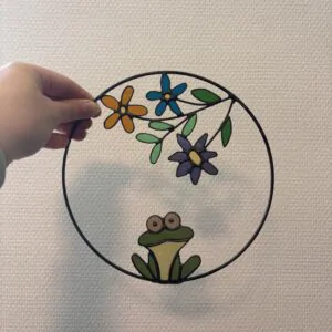 Glas in lood hanger: een metalen ring met daarin een kikker en 3 bloemen met een takje.