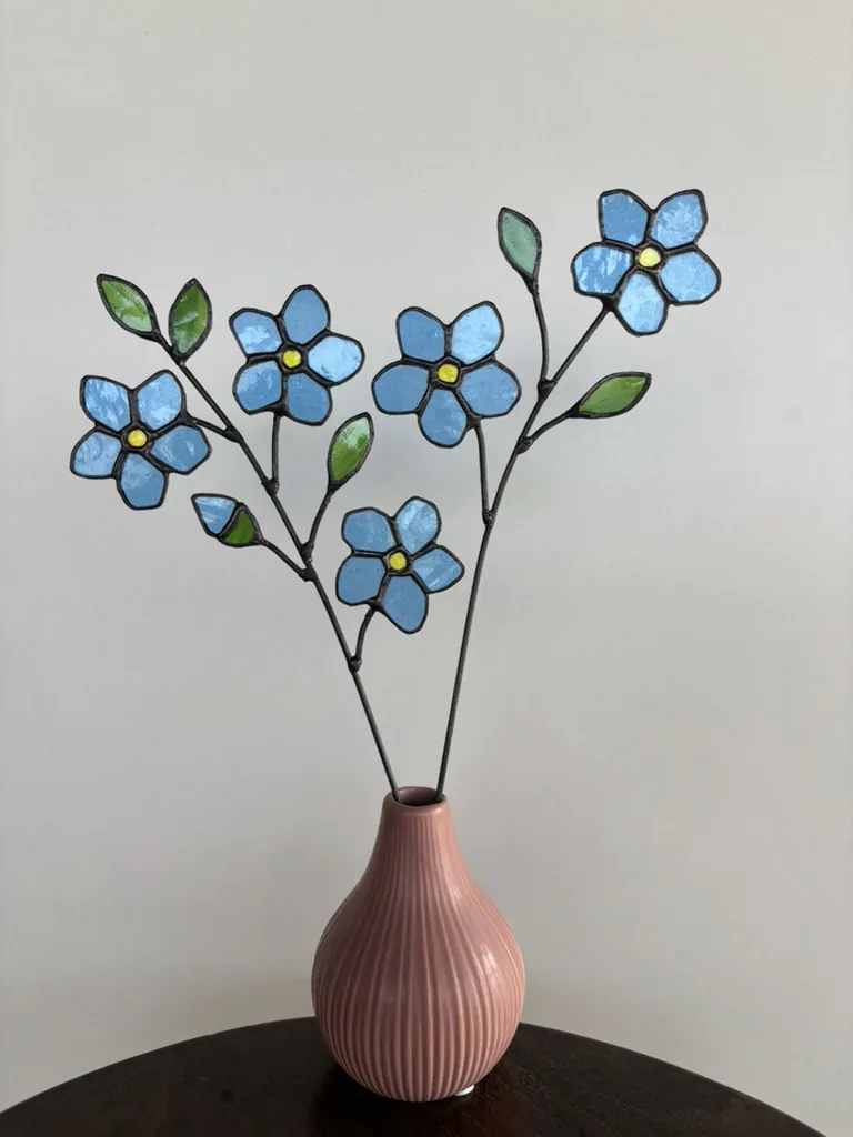 Tiffany glaskunst: sfeerfoto van een tak met 3 blauwe vergeet-mij-nietjes, 1 knop en 3 groene blaadjes en een tak met 2 blauwe vergeet-mij-nietjes, 2 groene blaadjes.